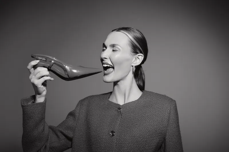 A smiling caucasian woman with light skin and slicked back hair holds a high heel shoe near her open mouth in a studio portrait. Concept of fashion, humor, and lifestyle.