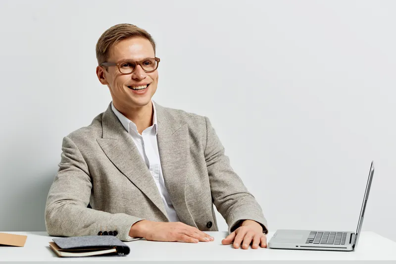 Smiling man in formal suit with glasses working at desk on laptop isolated on plain white background. Business professional, office, technology, success, confident, workspace, studio portrait.