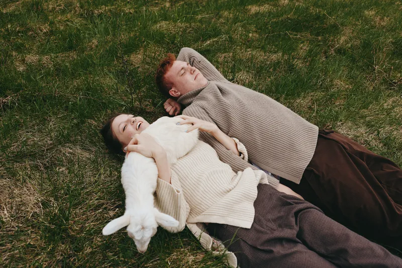 Young couple lies on grass enjoying a relaxed moment with a white baby goat in a casual lifestyle outdoor setting during autumn.