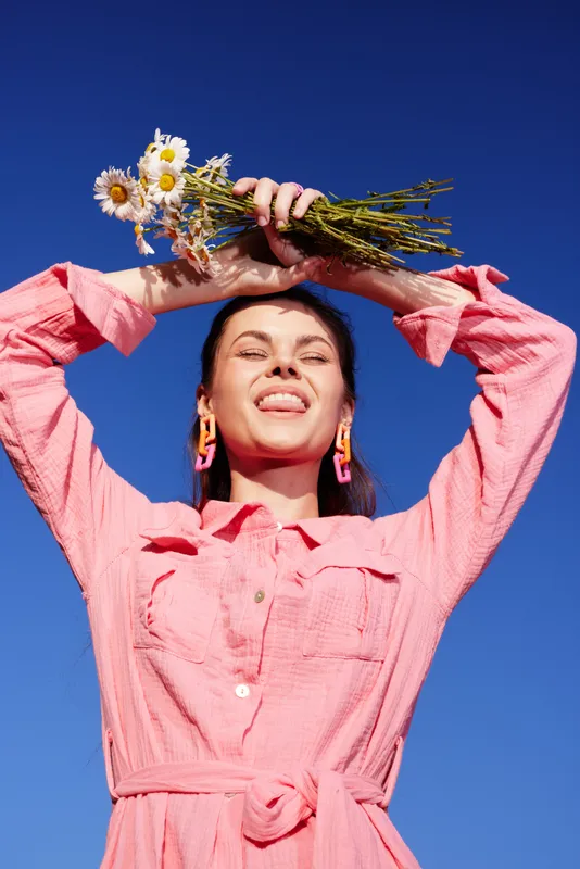 Smiling woman in a pink dress holding flowers against a blue sky, symbolizing joy and summer vibes Bright colors and joyful expression capture a cheerful mood