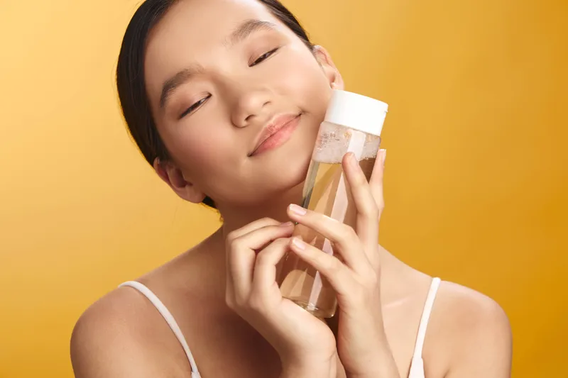 Asian woman smiles softly while embracing a skincare bottle, standing against a warm backdrop. Her calm expression conveys self care, wellness, and a modern beauty routine, creating an inviting and