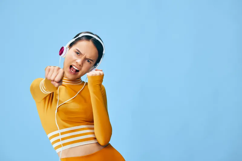 Energetic young woman fitness athlete in mustard sportswear and headphones, mid punch pose conveying strength and cardio workout intensity against a light blue background.