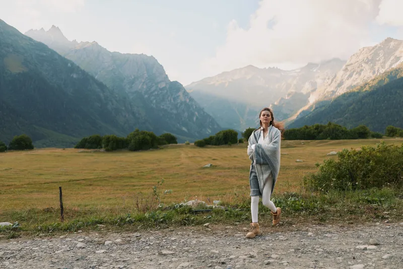 Young woman lifestyle walking on gravel path in mountain valley wrapped in warm blanket with cloudy sky and green grass background during autumn season.