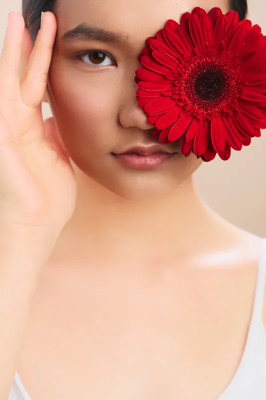 asian woman is seen with a vibrant red flower held beside her face in a soft portrait. her calm gaze and gentle expression convey elegance and poise.