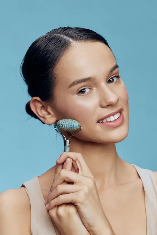 Young woman using a facial roller for skincare, promoting healthy beauty and self care rituals with a serene expression and a light blue background