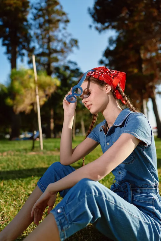 Lifestyle woman in modern boho-western denim street style sits outdoors with relaxed pose, wearing red bandana, film color look enhancing natural sunlight and shadows.