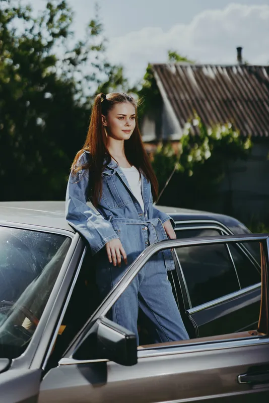 Young woman in denim outfit posing beside a retro car against a sunny backdrop, expressing confidence and style in a vibrant outdoor setting.
