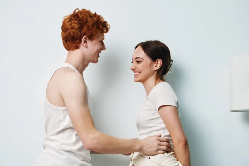 Young couple smiling and embracing indoors, man and woman facing each other with happy expressions in casual white clothing against plain background in romantic moment.
