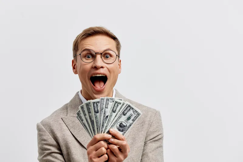 Jovial middle-aged man with glasses excitedly holding a fan of hundred-dollar bills, wearing a light gray suit, on a plain light background. Bright, cheerful, financial success concept, money, wealth,