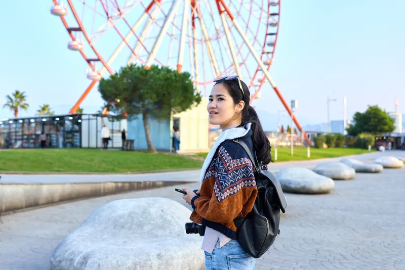 Traveler woman with camera at a park beside a ferris wheel, outdoor leisure setting with backpack, daylight glow, casual vibe, showcasing exploration and urban scenery