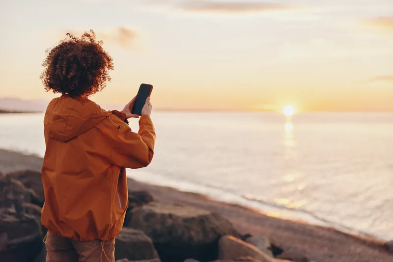 Woman lifestyle, smartphone, sunset, ocean, seashore, standing outdoors, orange jacket, curly hair woman takes photo by calm waves, warm golden hour background, travel inspiration