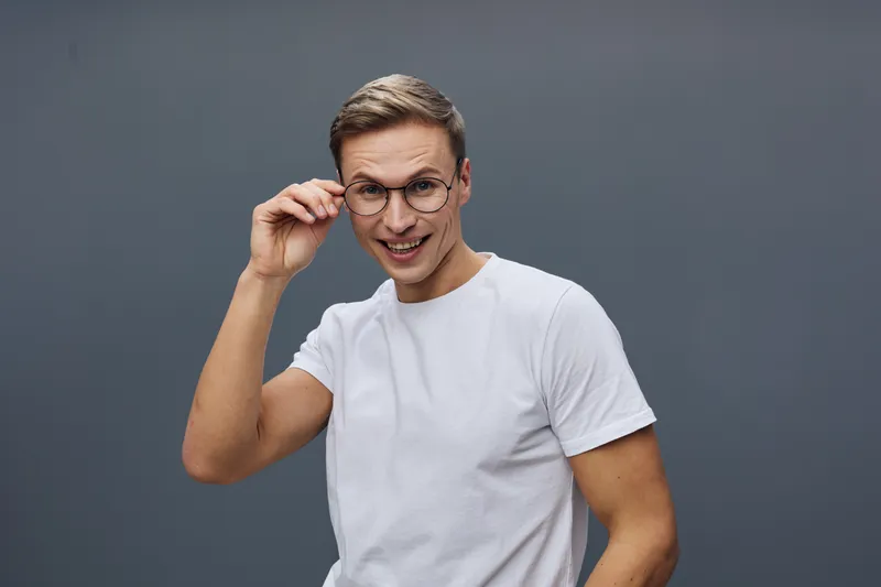 Smiling man with glasses, casual white t-shirt, playful expression, indoor studio portrait, friendly mood, young adult, light gray background, lifestyle photography.