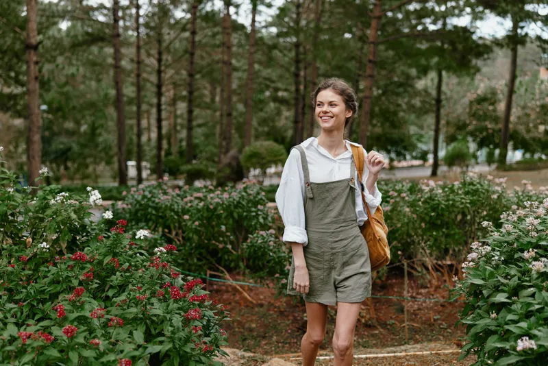 Young woman enjoying a sunny day in a lush garden, wearing casual overalls and smiling while carrying a bag, surrounded by vibrant flowers and greenery
