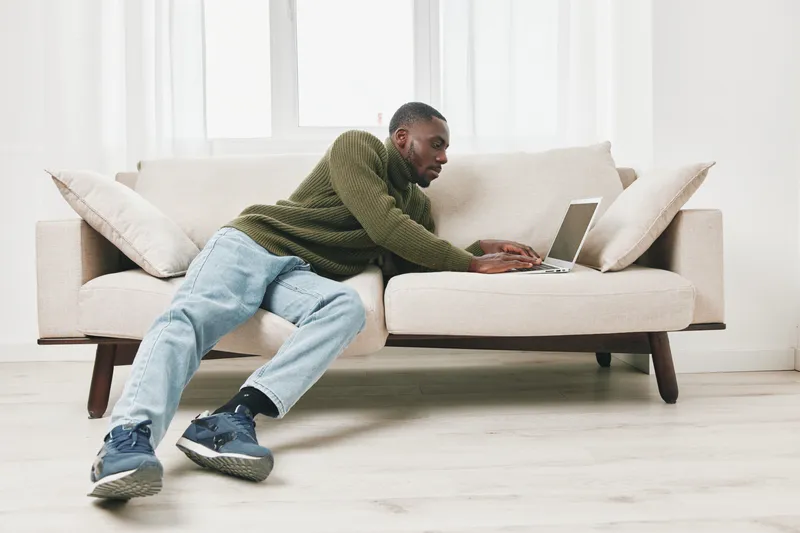 Young Black man working from home on a laptop while lounging on a stylish beige sofa, wearing a green sweater and relaxed jeans, creating a comfortable and modern atmosphere