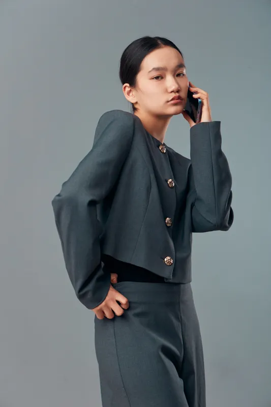 Young woman in stylish gray suit talks on smartphone posing confidently against plain background. Fashion model with dark hair and serious expression in studio portrait.