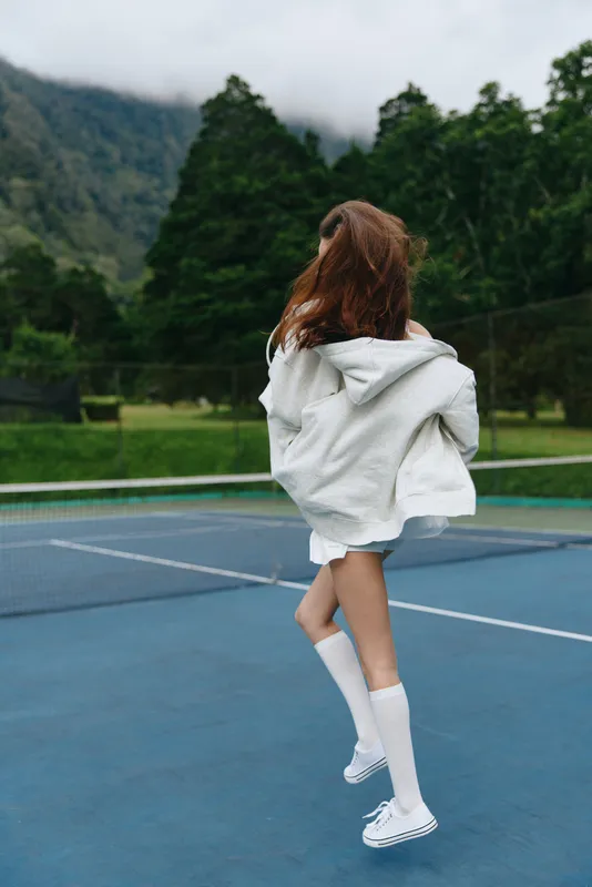 Young woman enjoying a carefree moment on a tennis court, wearing casual athletic attire, with flowing hair and a joyful expression, surrounded by nature