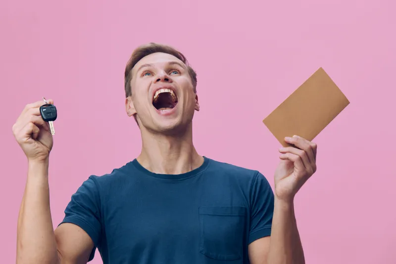 Happy young man with short brown hair, wearing casual blue t-shirt, holding car key and brown envelope, excited surprised expression, isolated on pink background, concept of success and achievement, l