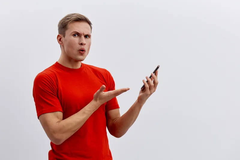 young caucasian man wearing red t-shirt holding smartphone confused speaking digital detox concept isolated on gray background