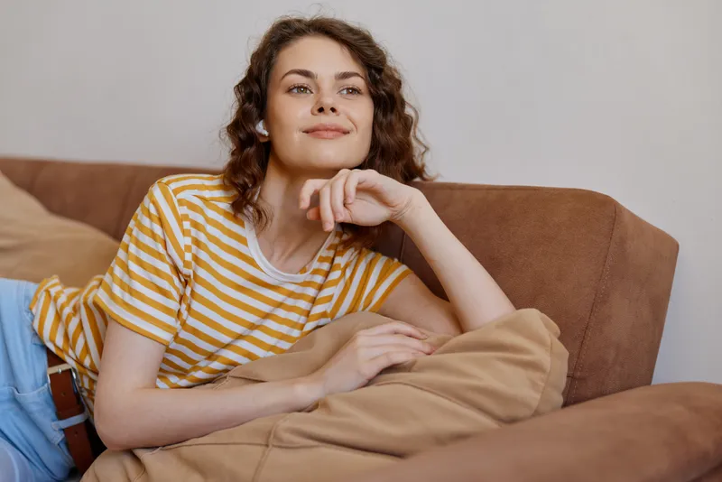 A relaxed woman on a sofa at home, wearing a striped yellow shirt, rests her chin on her hand. Concept for comfort, casual mood, cozy living room moment.