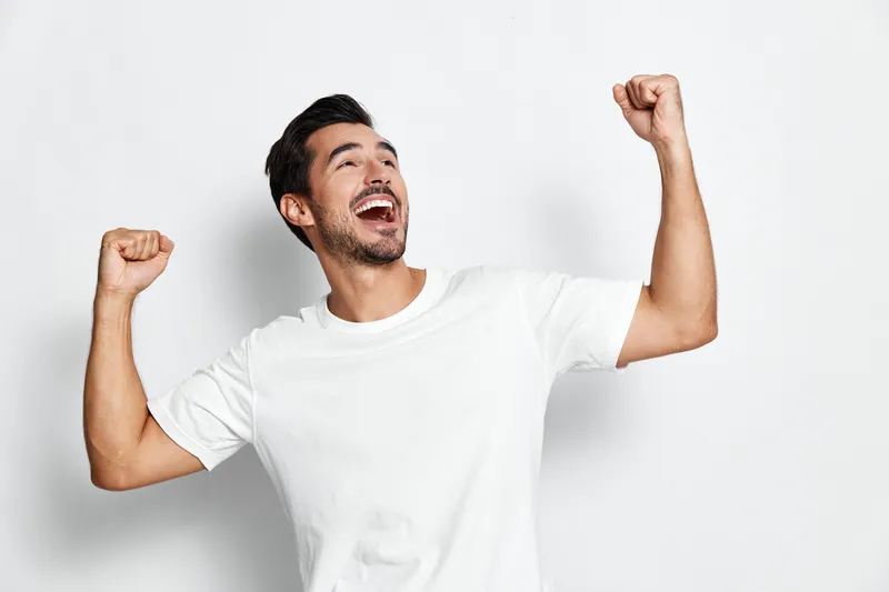 Full body joyful happy young man with short dark hair wearing a white t-shirt, celebrating success, raising hands in excitement, isolated on plain solid gray background.