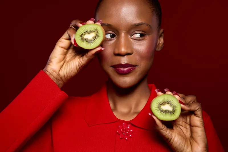 African woman with dark skin holds two halves of kiwi fruit near her face against red background. Concept of healthy lifestyle, natural beauty, fresh fruit, and skincare wellness.