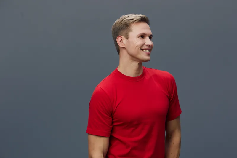 Smiling young man with blond hair wearing red t-shirt looking to the side against plain gray background studio portrait. People lifestyle concept