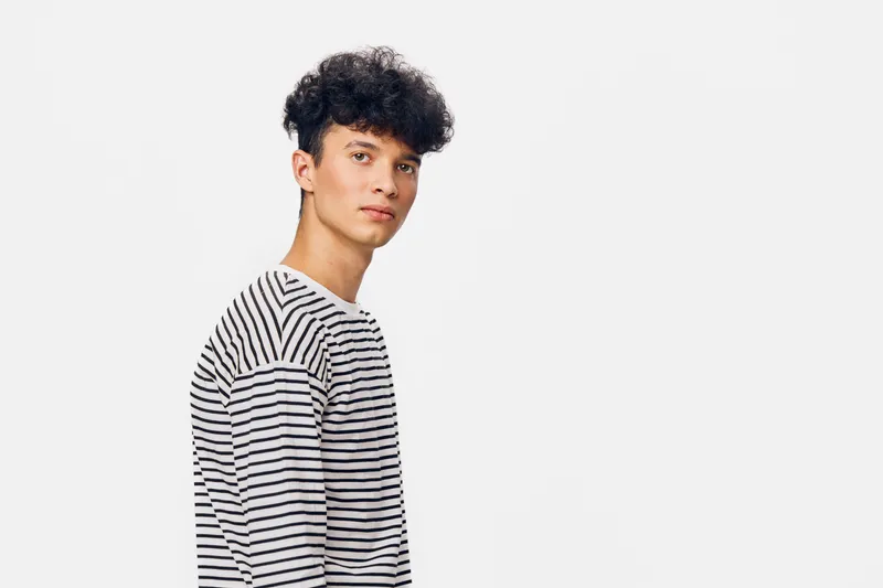 Young man with curly hair wearing striped shirt stands against white background, looking confident and thoughtful in casual style portrait.