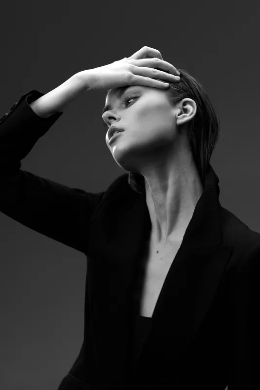 Black and white portrait of a thoughtful woman in a blazer with hand on forehead, expressing elegance and introspection.
