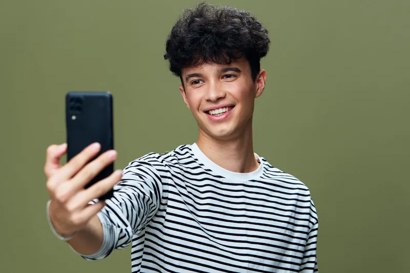 Young man taking selfie with smartphone, smiling joyfully wearing striped shirt against green background, casual lifestyle and modern technology concept.