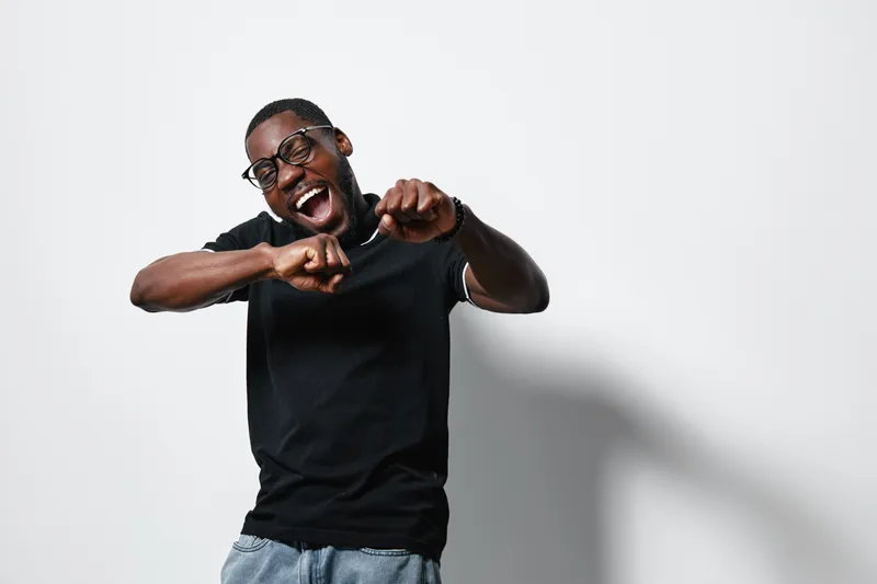 Black man with dark skin wearing glasses and black t shirt dancing and smiling on white background with shadow. Casual style and joyful expression in studio.