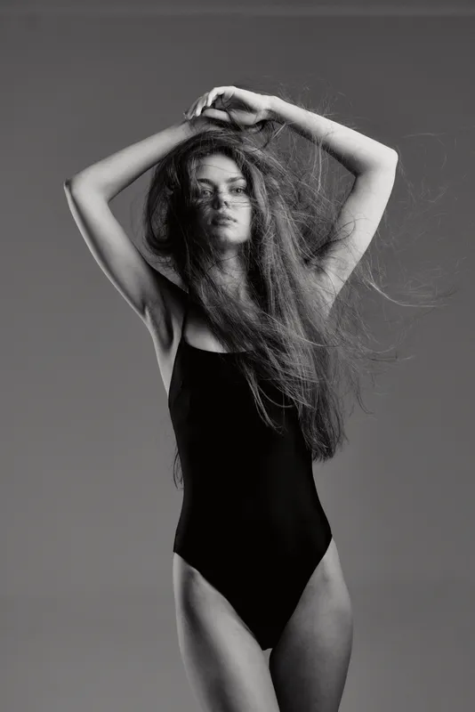 Black and white portrait of a young woman with long hair wearing a black bodysuit, posing with arms raised against a plain background.