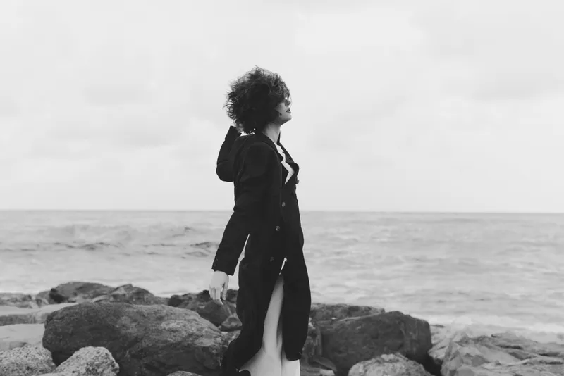Woman in a long coat stands on a rocky shore by the sea, wind in her hair, looking toward the horizon in a moody monochrome landscape
