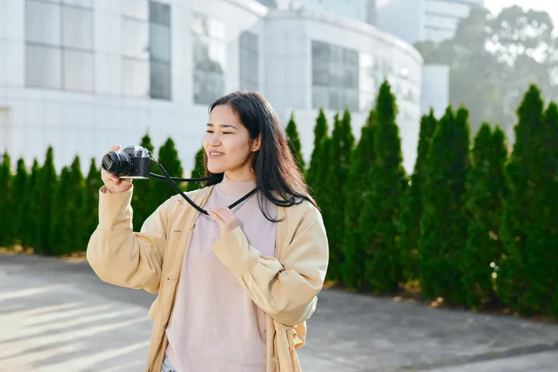Camera in hand, a woman photographer shoots outdoors along a modern building complex, wearing a beige jacket and soft sweater, capturing candid scenes with confident style and focus.