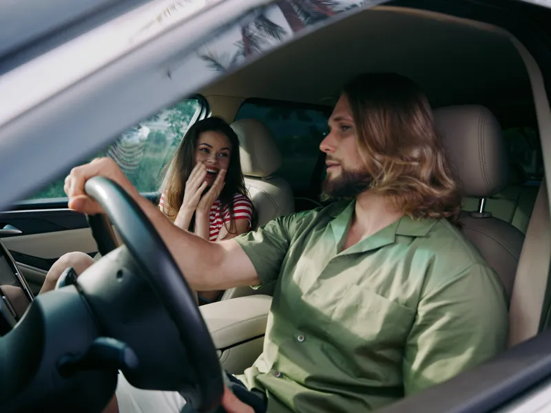 A man with shoulder length hair drives a car while a surprised woman in the passenger seat giggles, capturing a candid and light hearted moment inside a modern vehicle.