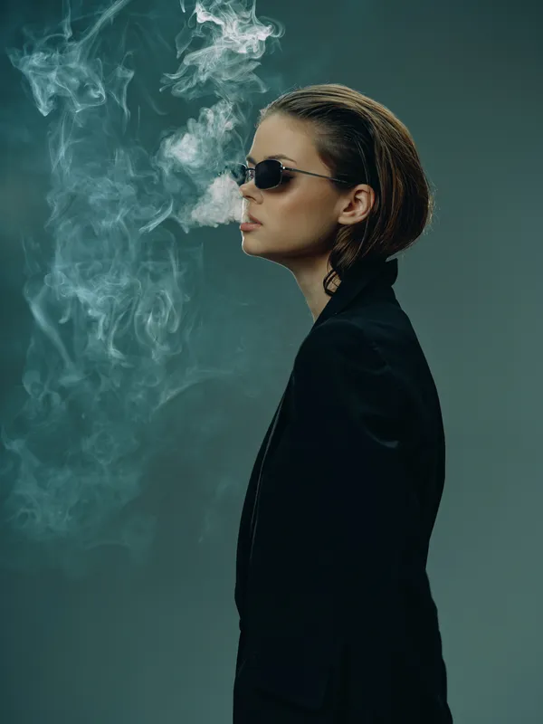 Side view of a stylish woman wearing sunglasses, exhaling smoke, dressed in black with moody lighting and a dark background.