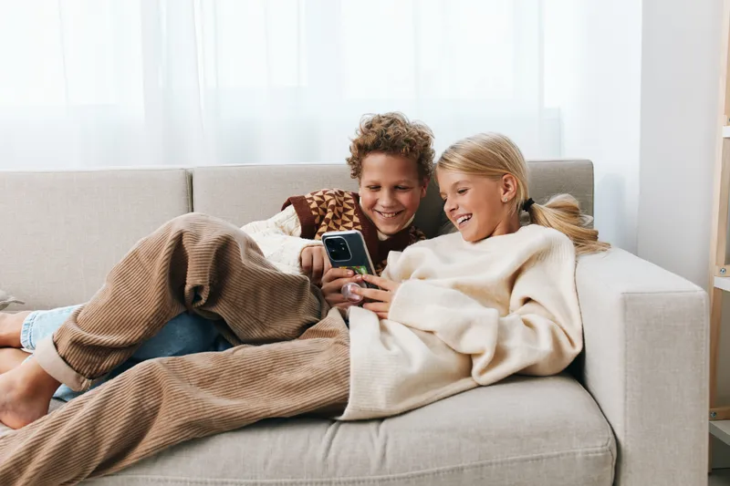 Authentic candid moment shows children resting on a sofa, exploring a phone in a cozy living space, blending play and daily school life with ease.