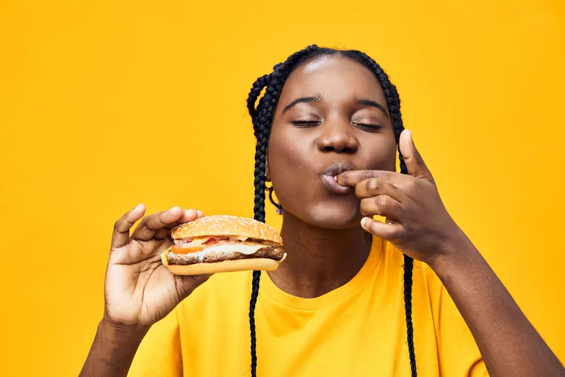 Young African woman enjoying a delicious burger, showcasing happiness and satisfaction against a vibrant yellow background, perfect for healthy eating concepts