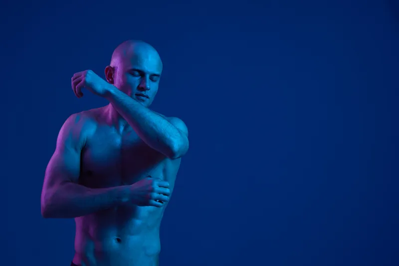 A man poses gracefully against a vivid blue backdrop, showcasing his toned physique in a dynamic and expressive manner. The light enhances the shapes and shadows.