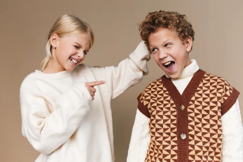 Two children share a playful moment as a girl in a white sweater taps the boy’s head, both smiling brightly, creating a joyful, candid scene against a warm neutral studio backdrop.