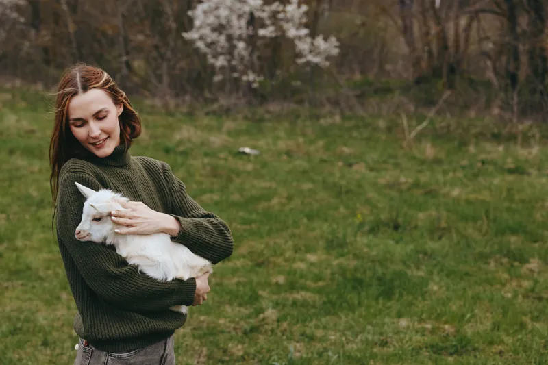 Young woman with long hair gently holds a white baby goat in a green field during spring, enjoying a peaceful moment in nature and a cozy knitted sweater.