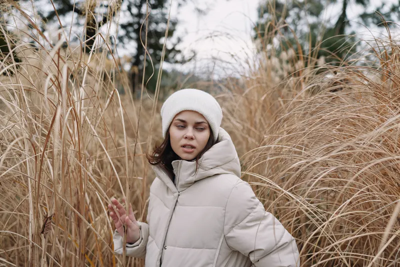 Winter girl in a puffy coat standing in tall grass outdoors, calm expression, soft light, nature scene, cold air, cozy apparel. Soft breeze, muted colors. cozy.
