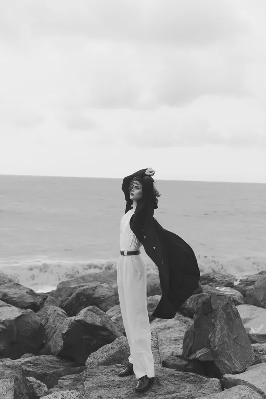 Monochrome image of a stylish woman on rocky shore, cape blowing in wind, sea behind, elegant dress and relaxed pose by the coast