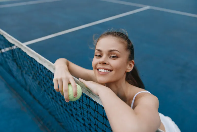 Young woman smiling while holding a tennis ball on a blue court, showcasing athleticism and joy in a summer sports lifestyle