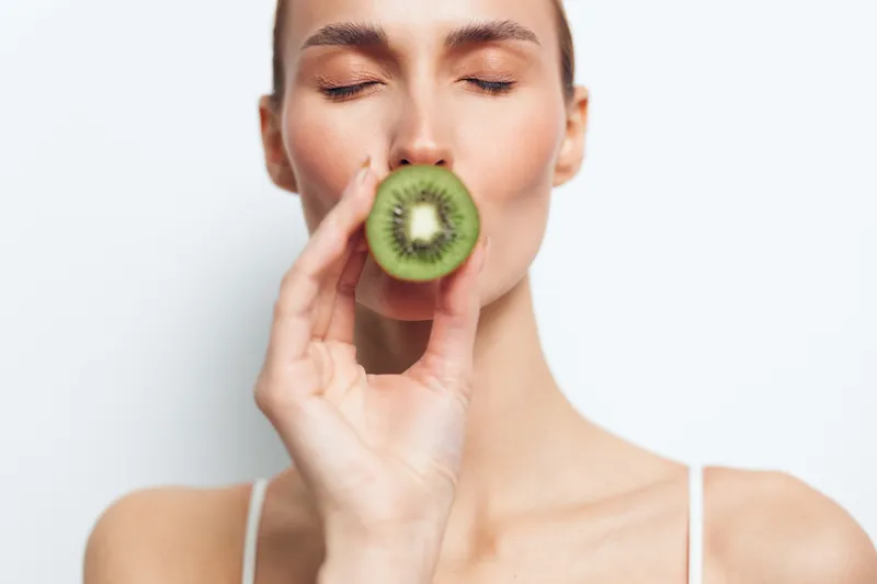 Closeup of a woman holding a kiwi to her lips, fresh fruit moment, natural lighting, clear skin, relaxed expression, vibrant green tones, simple lifestyle portrait, serene mood