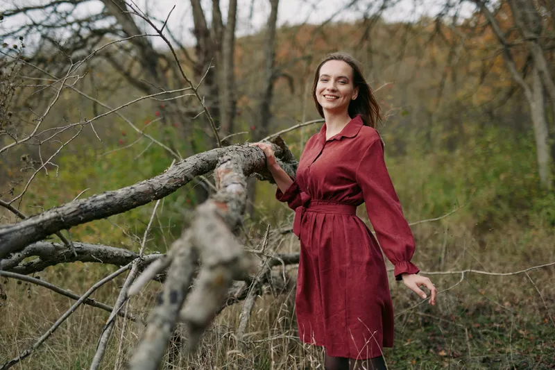 Smiling woman in a red dress stands by a fallen tree in a woodland clearing, captured as a genuine candid portrait emphasizing natural expression, honest emotion and real presence.