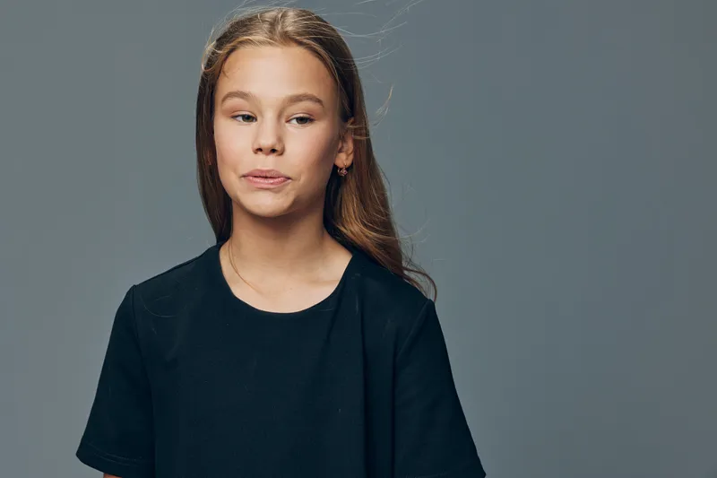 Teenage girl with long hair wearing black shirt standing against gray background looking sideways with thoughtful expression. Portrait of young female in casual clothing studio shot.