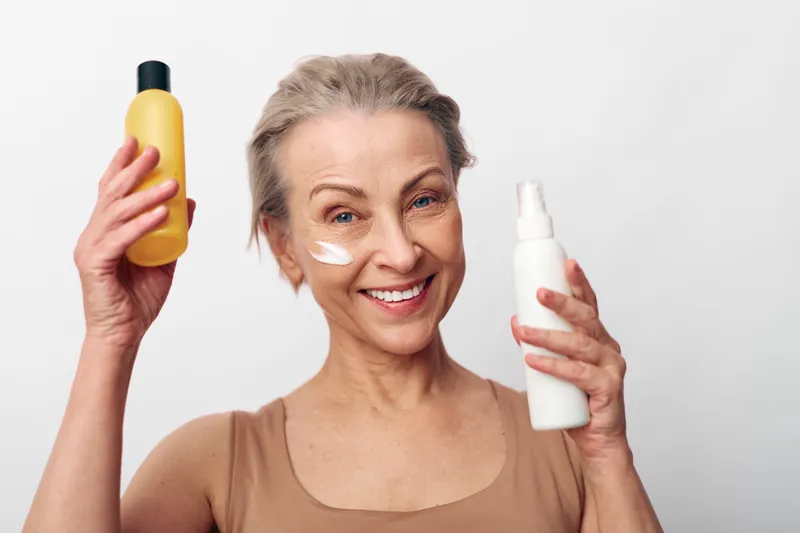 Smiling mature woman with natural skin holds two skincare bottles applying cream on cheek. Concept of skincare routine, beauty care, anti aging, wellness, and self confidence in studio.