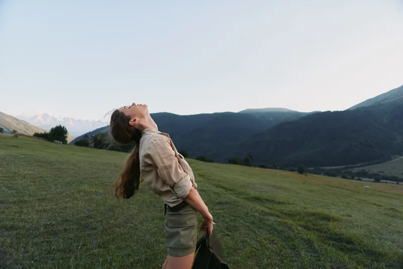 Young woman with long hair leans back with closed eyes enjoying fresh air in a green mountain meadow during a clear day.