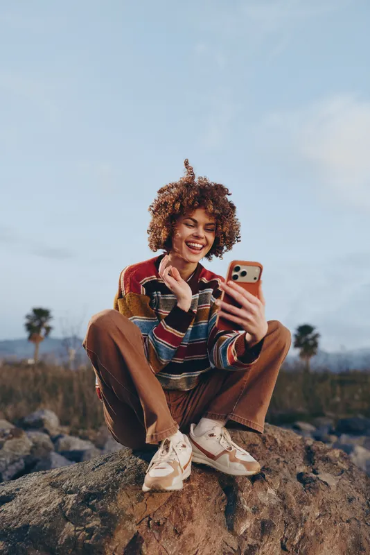 Woman lifestyle outdoors selfie smartphone smiling curly hair rock casual, young woman sitting on stone taking photo with phone, striped sweater and jeans, warm evening light, natural landscape