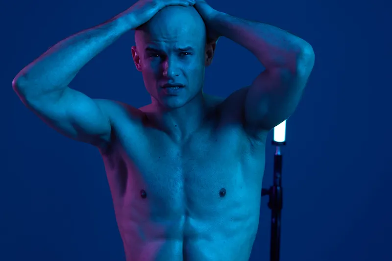 Intense man in blue light, expressing emotion with bare torso and hands on head, showcasing strength and vulnerability in a striking composition.